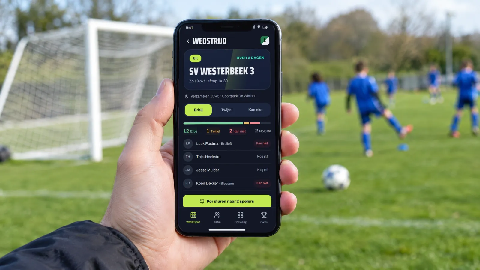 Telefoon met de Basiself-app die laat zien wie er bij de wedstrijd is, voor een veld met jeugdspelers